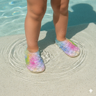 Child's feet wearing Finny Shoes colorful mermaid scale sandals on a sandy beach.